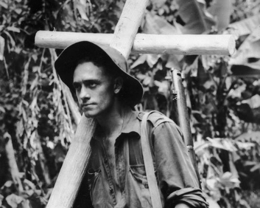 An Australian infantryman carries a cross hewn from rough forest timber to a forward position to mark the burial place of a fallen comrade (AWM 018176).